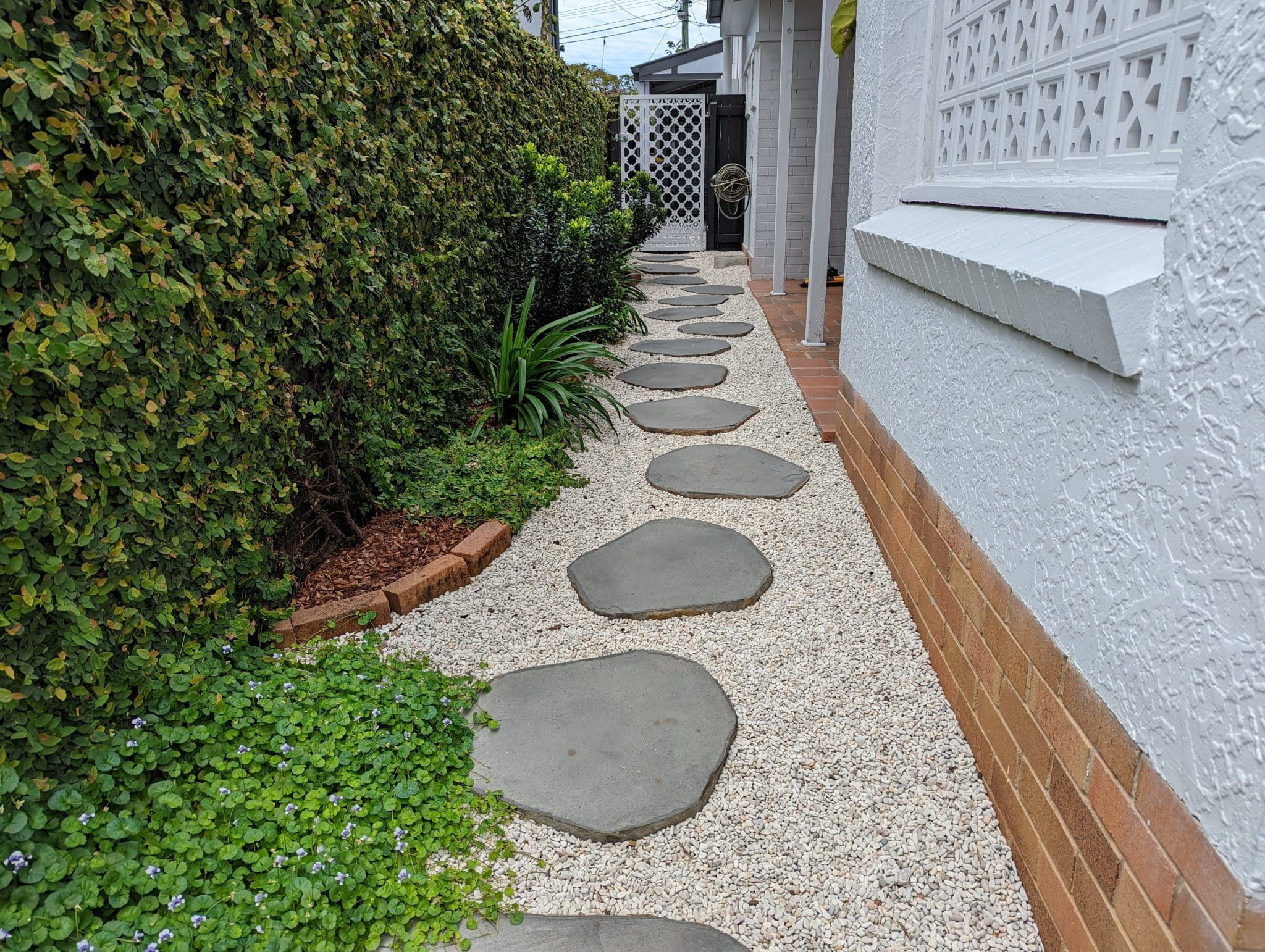 Natural stone stepping stones laid in a modern garden path bordered by lush greenery and white gravel beside a white rendered house.