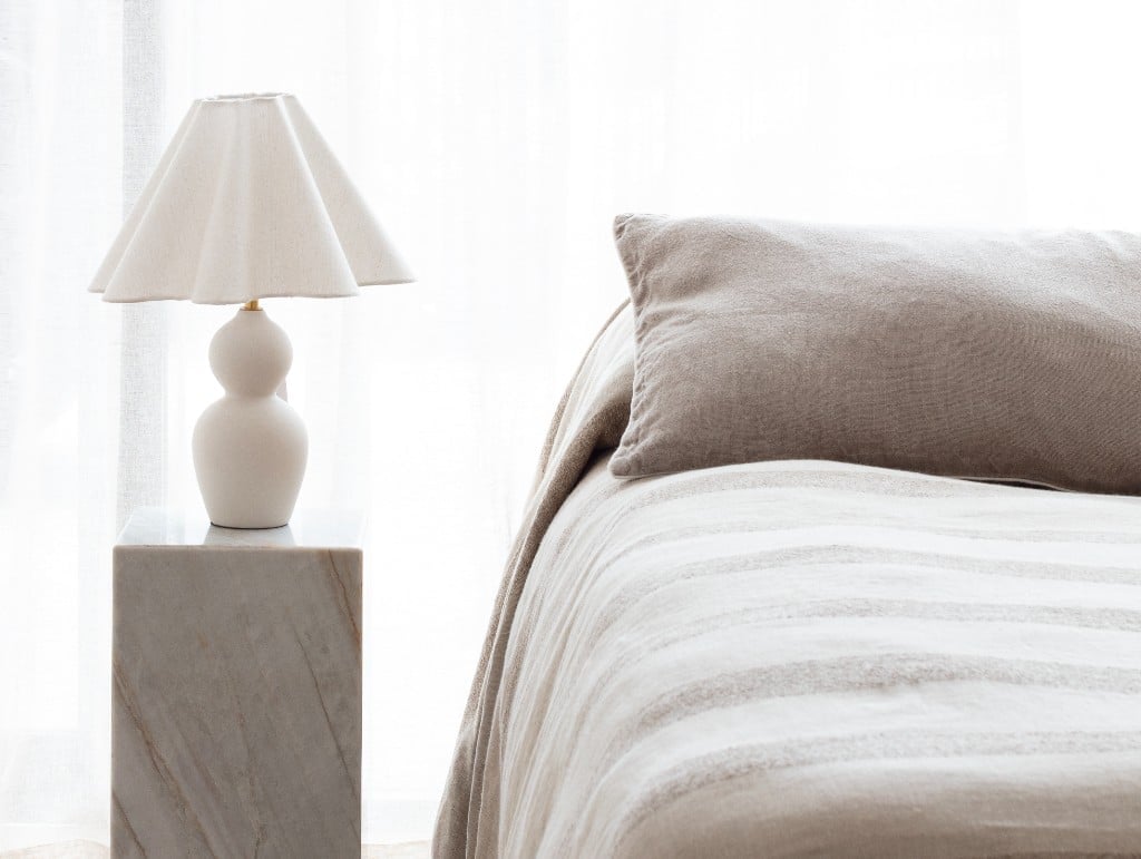 Bedroom interior featuring In The Sac natural linen bedding in soft neutral tones, styled with a Paola & Joy wavy lamp on a stone plinth for a calm, minimalist aesthetic.