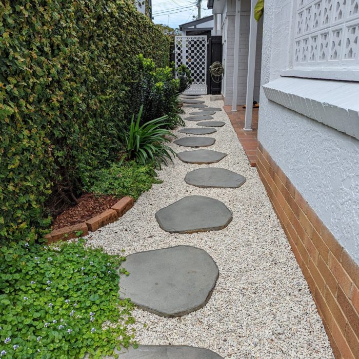 Natural stone stepping stones laid in a modern garden path bordered by lush greenery and white gravel beside a white rendered house.